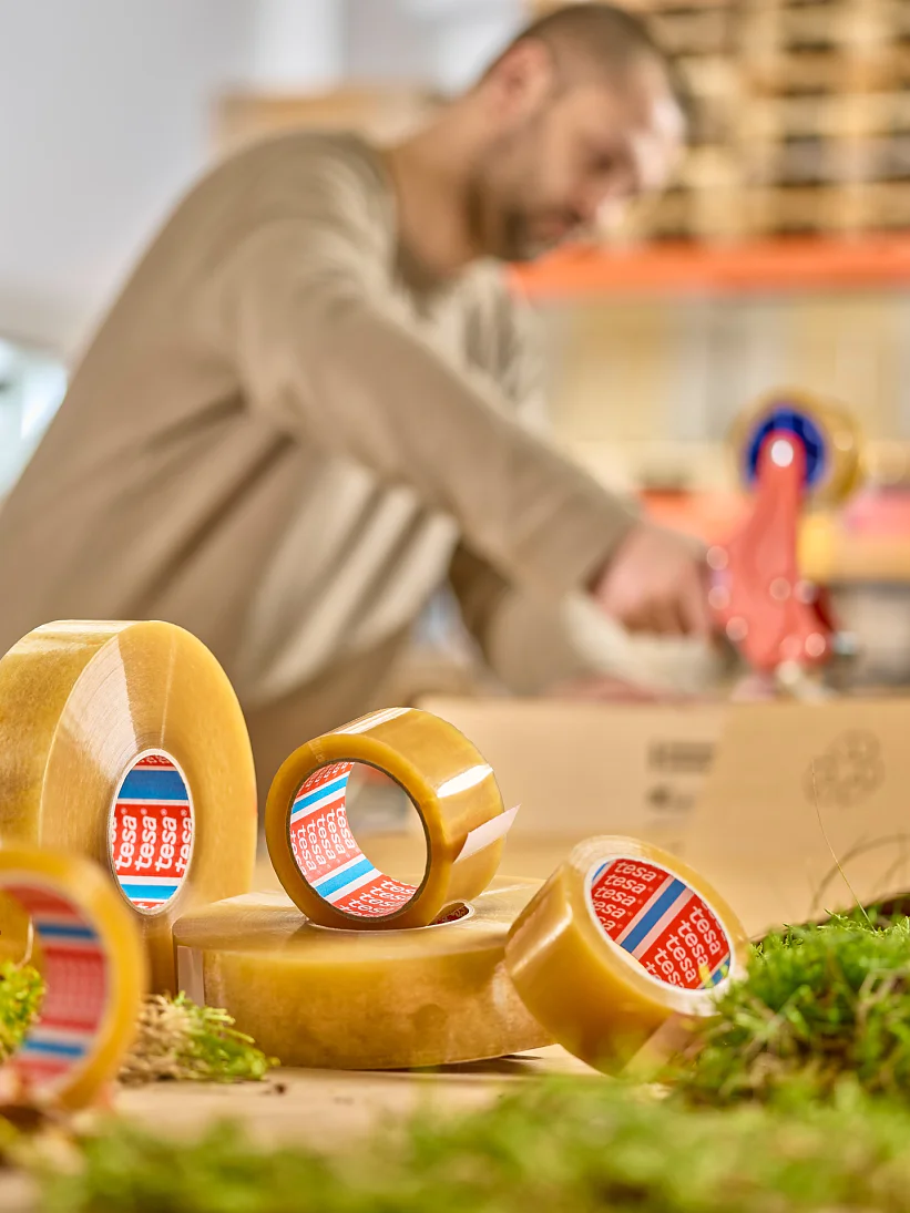 A person in a beige sweater is using a tesa tape dispenser in a warehouse setting. Several rolls of tesa tape are in the foreground, surrounded by patches of greenery. Shelves with boxes and pallets are visible in the background. (This text has been generated by AI)