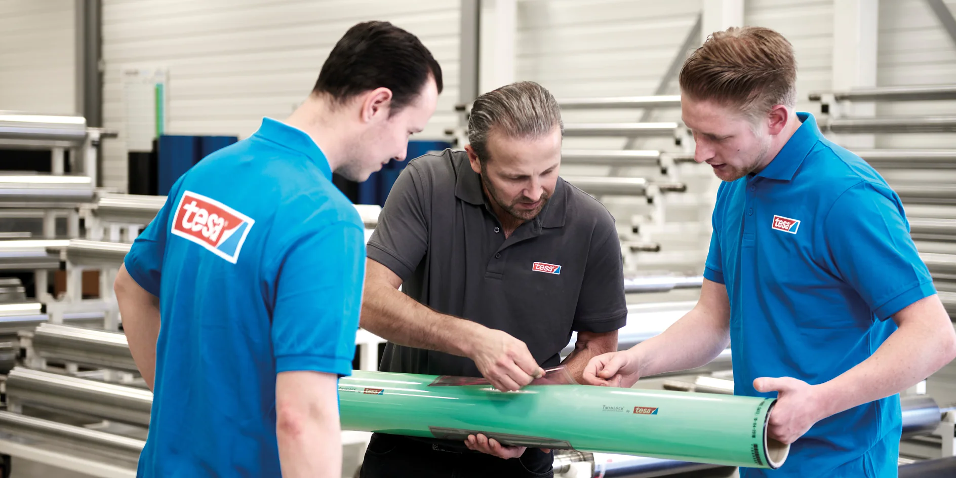 Three men in a factory setting examine a large green cylindrical object. Two men wear blue shirts with red logos, and the third, in the center, wears a gray shirt with the same logo. Metal pipes and machinery are visible in the background. (This text has been generated by AI)
