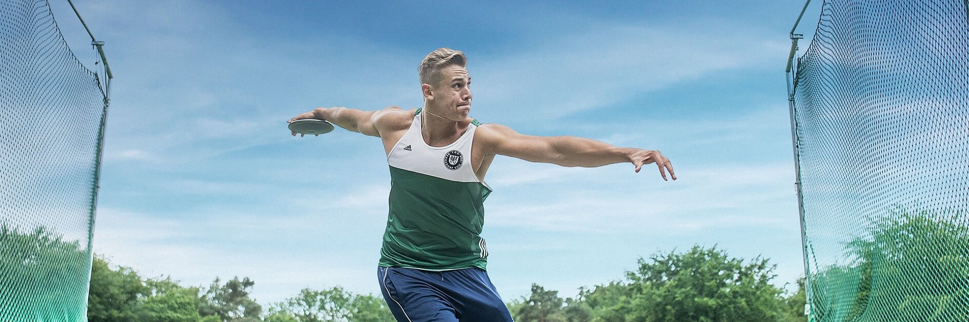 A male athlete in athletic attire is in mid-action, throwing a discus. He stands on a grassy field surrounded by a metal cage, with trees in the background under a clear blue sky.