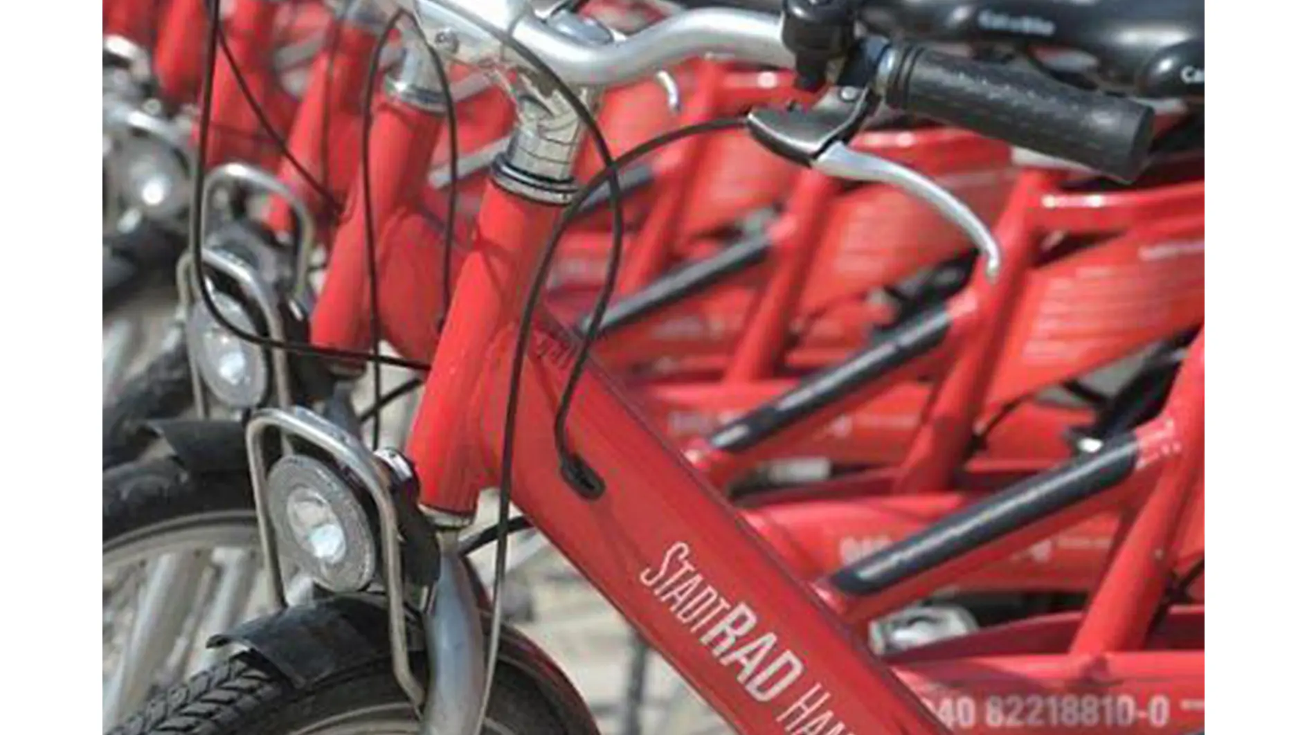 Close-up of red rental bicycles lined up in a row. The front wheel and part of the frame with the text StadtRAD Hamburg are visible. Each bike is equipped with a headlight and a cable lock. The handlebars and fenders are also in view. (This text has been generated by AI)