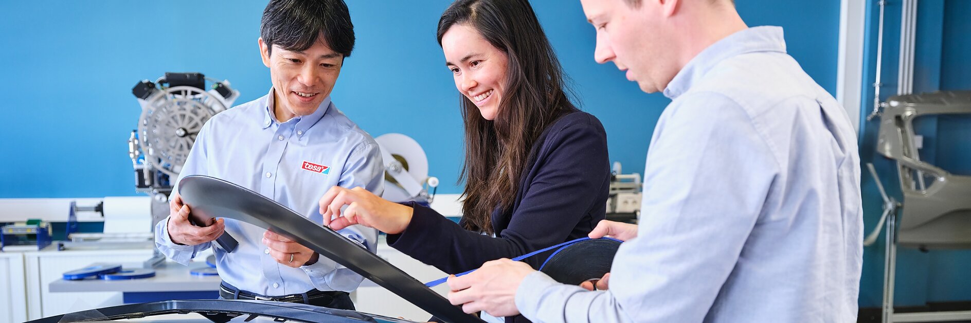 Three people examine automotive components on a table. The parts include strips and paneling secured with tesa tape. They are in a brightly lit room with a blue wall and additional components visible in the background.