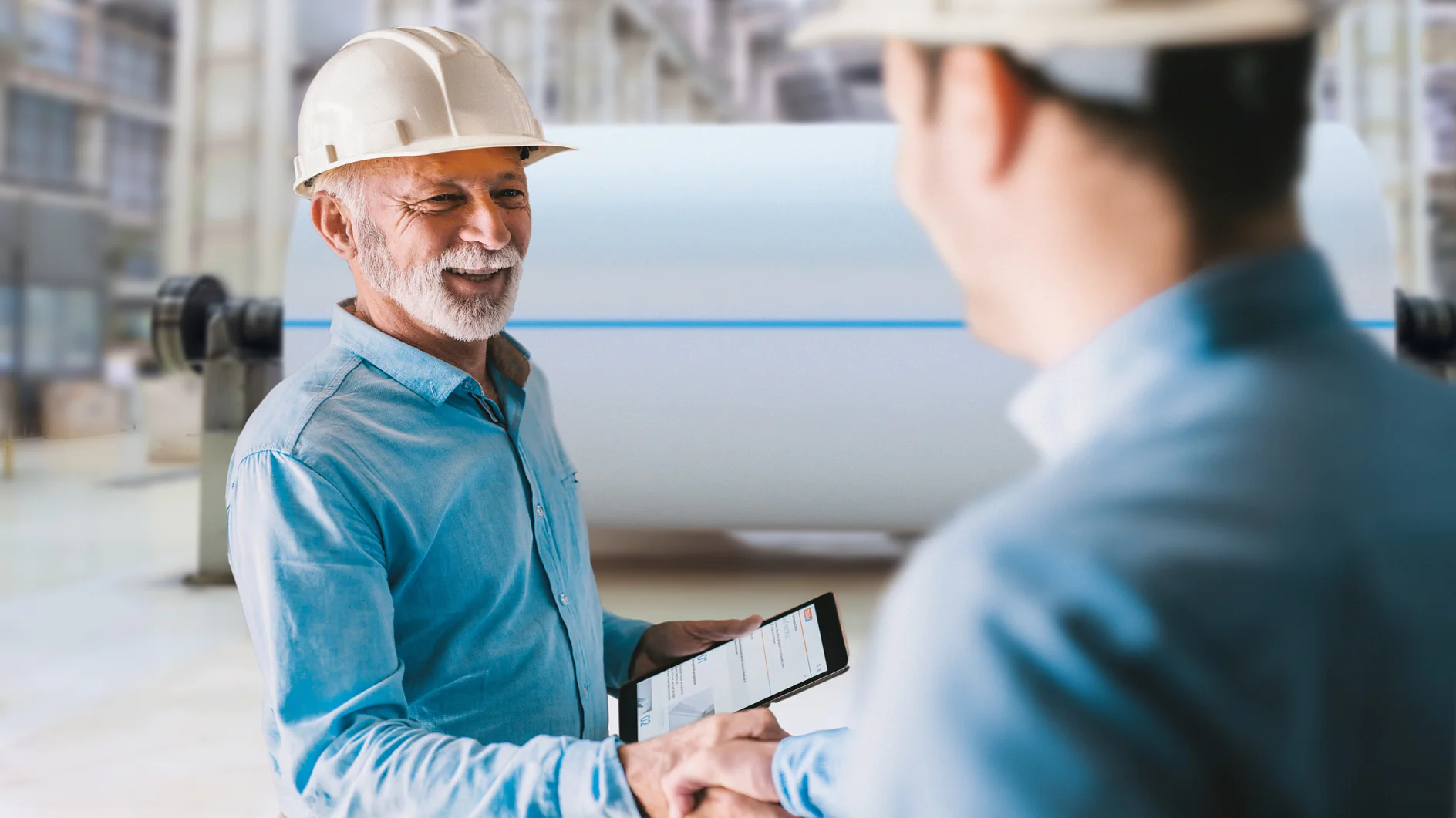 Two men wearing safety helmets shake hands in an industrial setting. One holds a tablet displaying graphs. Large machinery is blurred in the background, suggesting a factory or manufacturing environment, where tesa tape might be commonly used for various applications. (This text has been generated by AI)