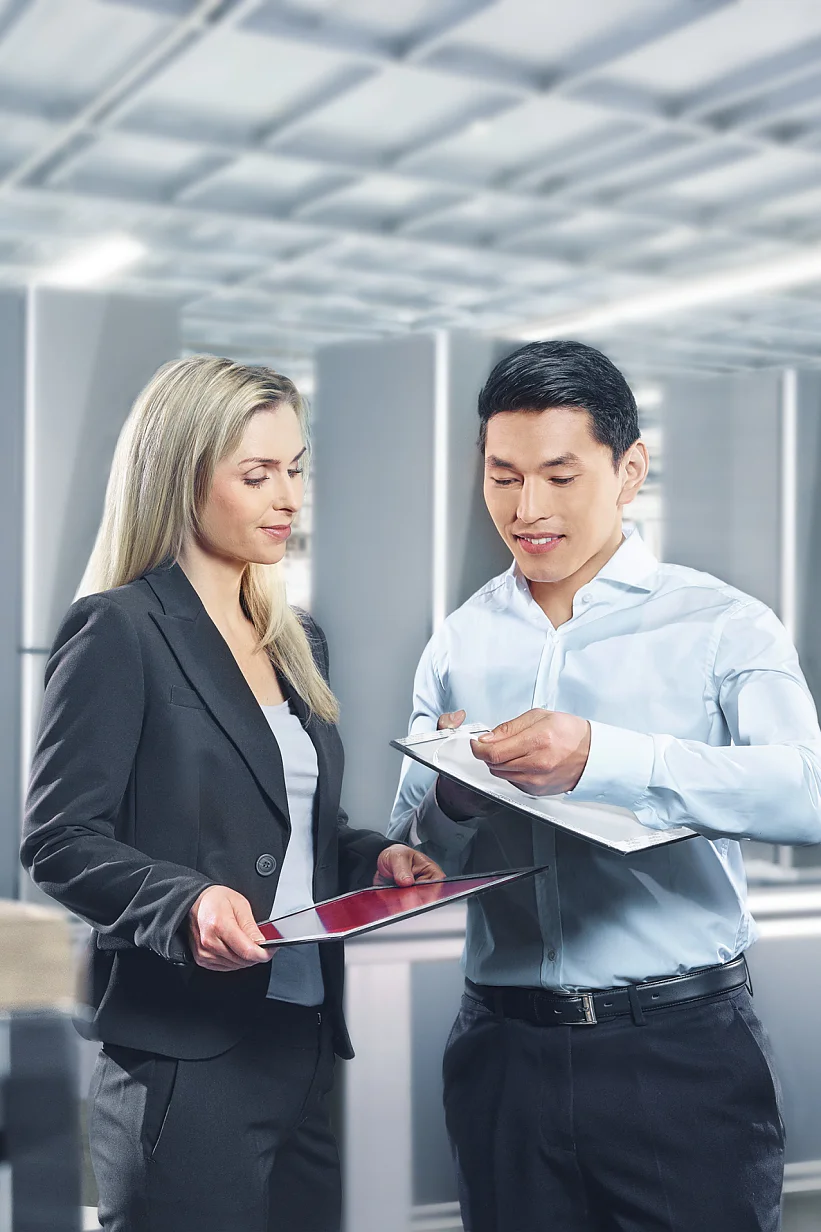 Appliances Gespiegelt A man and a woman are discussing documents in a bright industrial setting. The woman holds a tablet, and the man holds a clipboard secured with tesa tape. They are both professionally dressed. Equipment and large metal structures are visible in the background. (This text has been generated by AI)
