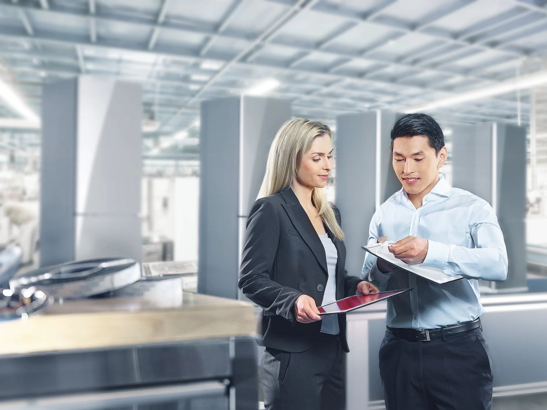 A man and a woman are discussing documents in a bright industrial setting. The woman holds a tablet, and the man holds a clipboard secured with tesa tape. They are both professionally dressed. Equipment and large metal structures are visible in the background. (This text has been generated by AI)