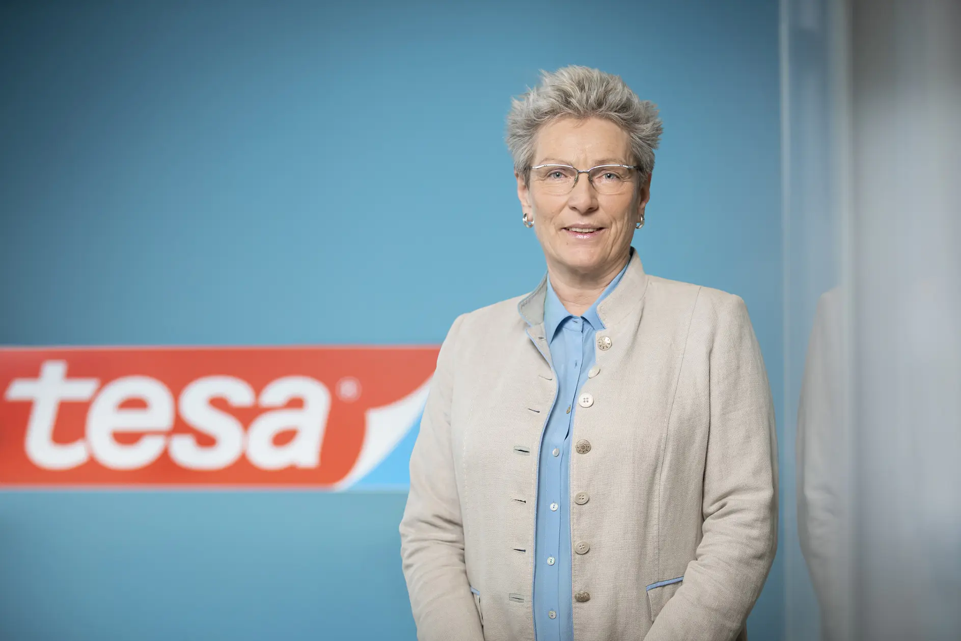 A person with short gray hair, glasses, and a beige blazer stands in front of a blue wall with a red and white logo that reads tesa tape.