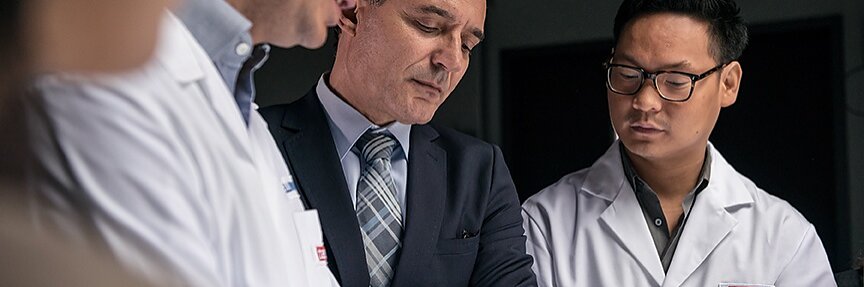 Four men are gathered, three wearing white lab coats and one in a suit. They appear to be discussing something of interest or examining an object outside the frame, possibly involving tesa tape. The setting looks like a professional or laboratory environment.