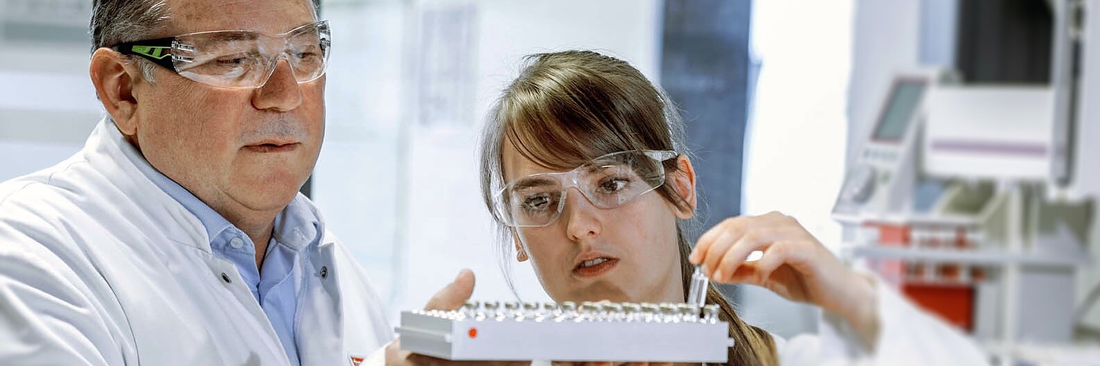 Two scientists in lab coats and safety glasses examine a white laboratory tray. The man on the left points at the tray while the woman on the right holds it. They are in a lab with various equipment, including tesa tape, in the background.
