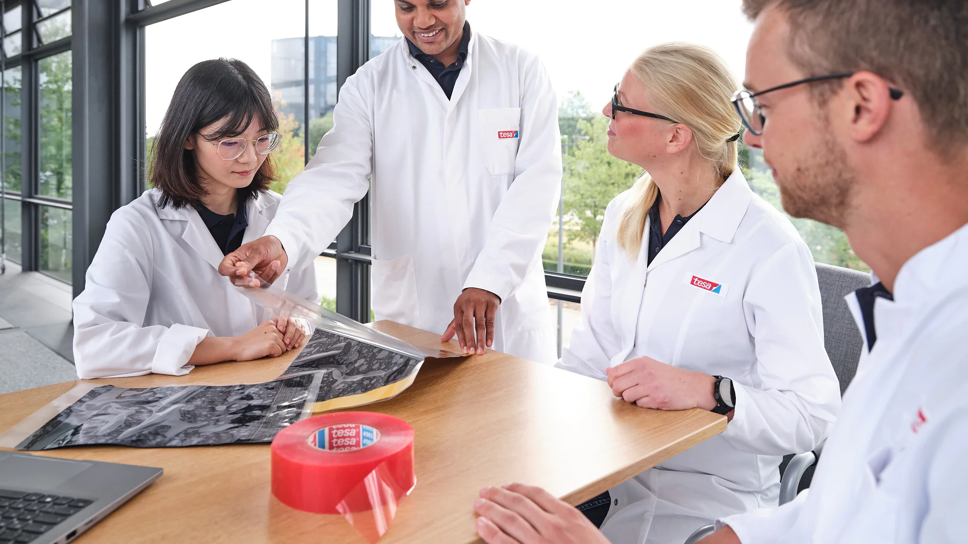 Four people in lab coats are gathered around a wooden table in a bright room with large windows. One person is showing a large sheet of material to the others. There is a roll of red tesa tape and a laptop on the table. (This text has been generated by AI)