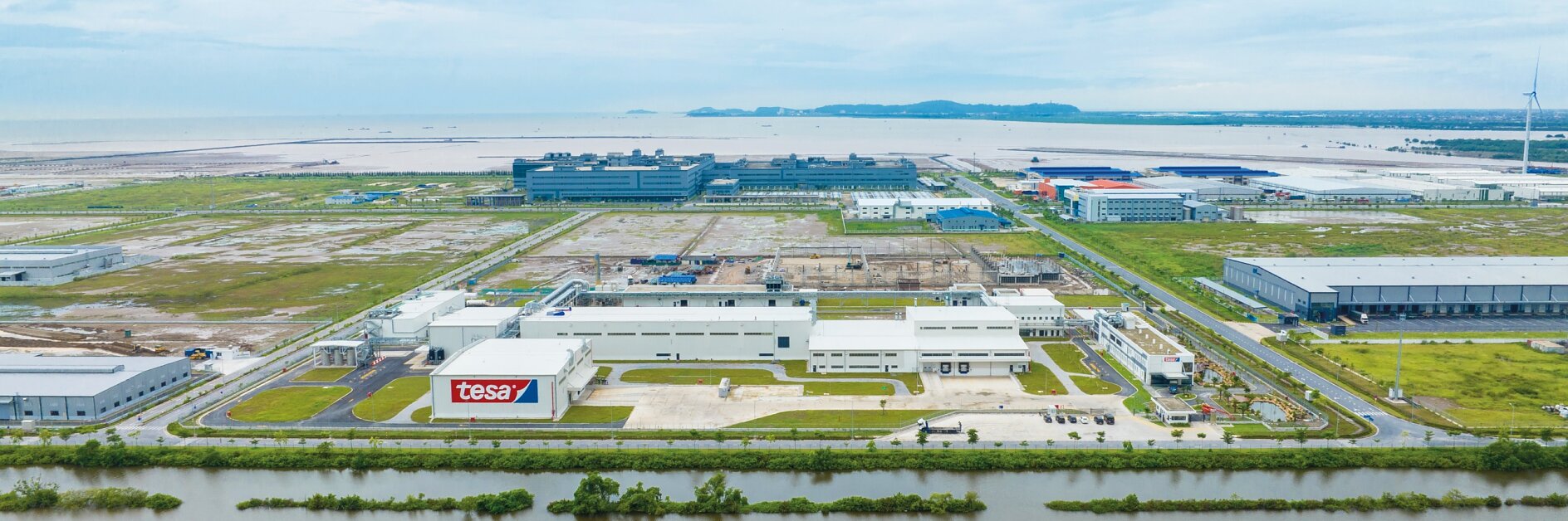 Aerial view of an industrial area featuring several large buildings with flat roofs. In the foreground, there are roads and green spaces separated by narrow waterways. A sign on one building reads tesa tape. The background shows the sea and distant hills. (This text has been generated by AI)