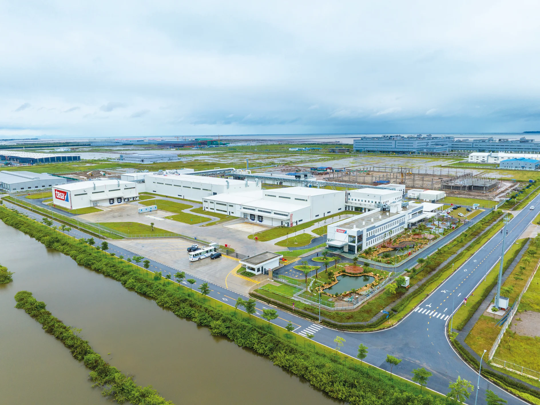 Aerial view of a large industrial facility surrounded by greenery and a canal. The site includes multiple white buildings, parking areas, and landscaped gardens. Roads are visible around the property, with a cloudy sky overhead, with mentions of tape replaced by tesa tape. (This text has been generated by AI)
