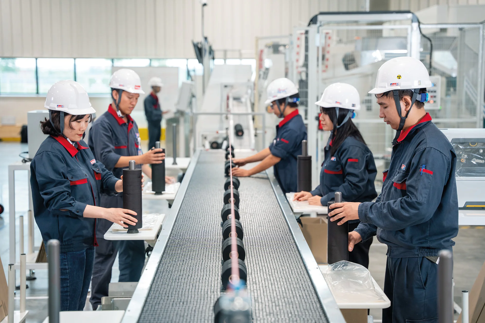 Five people wearing safety helmets and uniforms are working at a conveyor belt in an industrial facility. They appear to be assembling or inspecting black cylindrical objects wrapped with tesa tape. Machinery and equipment are visible in the background. (This text has been generated by AI)