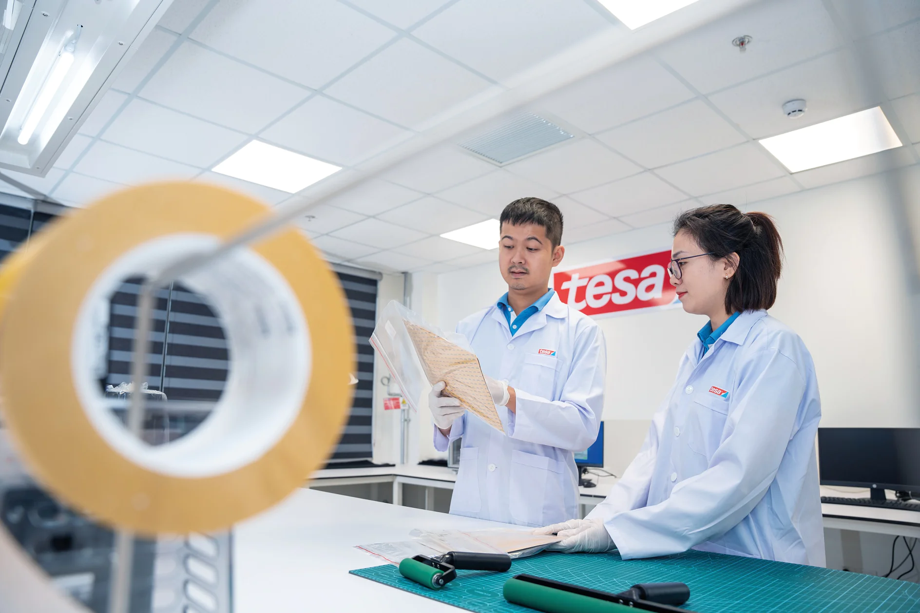 Two people in lab coats examine a large sheet of material in a laboratory setting. In the foreground, a roll of tesa tape is visible. The background has a sign with partially visible text. The room is well-lit with a modern design. (This text has been generated by AI)