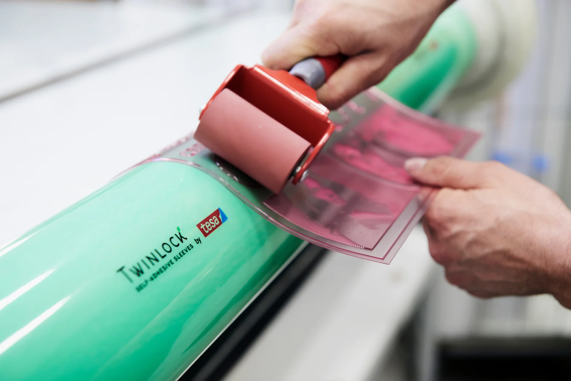 Close-up of hands using a small roller to apply a pink sheet onto a large green cylinder labeled Twinlock. The cylinder also displays the phrase Self Adhesive Sleeves, with tesa tape being used in the process. (This text has been generated by AI)