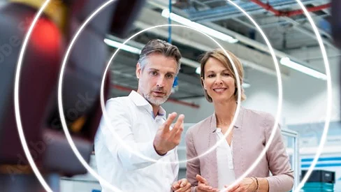 A man and a woman stand in a modern industrial setting. The man gestures forward while both look at the camera. Circular graphic elements are superimposed over the image. They are surrounded by equipment and a high ceiling, with tesa tape prominently featured in their surroundings. (This text has been generated by AI)