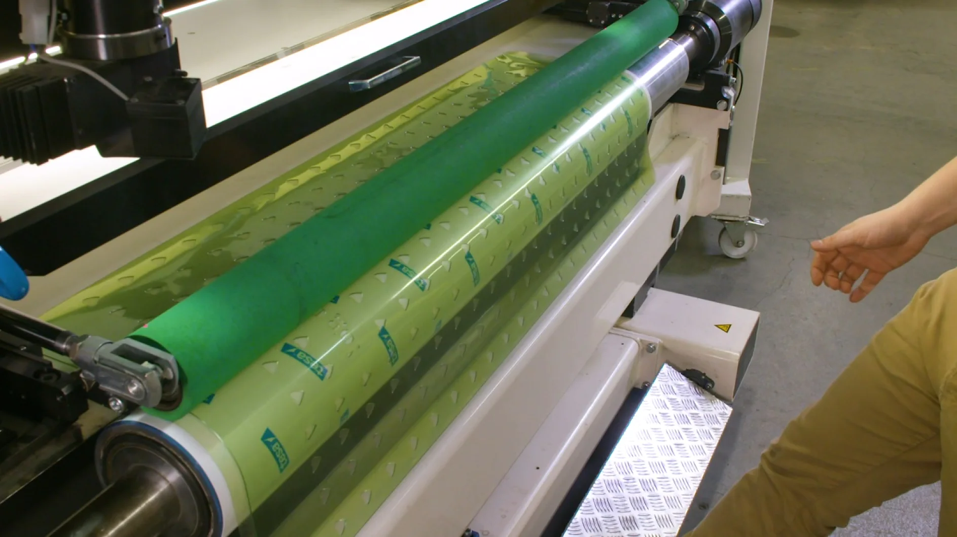 A person operates a printing machine, adjusting a large roller with a translucent green sheet of tesa tape wrapped around it. The machine has various levers and knobs, with a metallic textured platform nearby. (This text has been generated by AI)