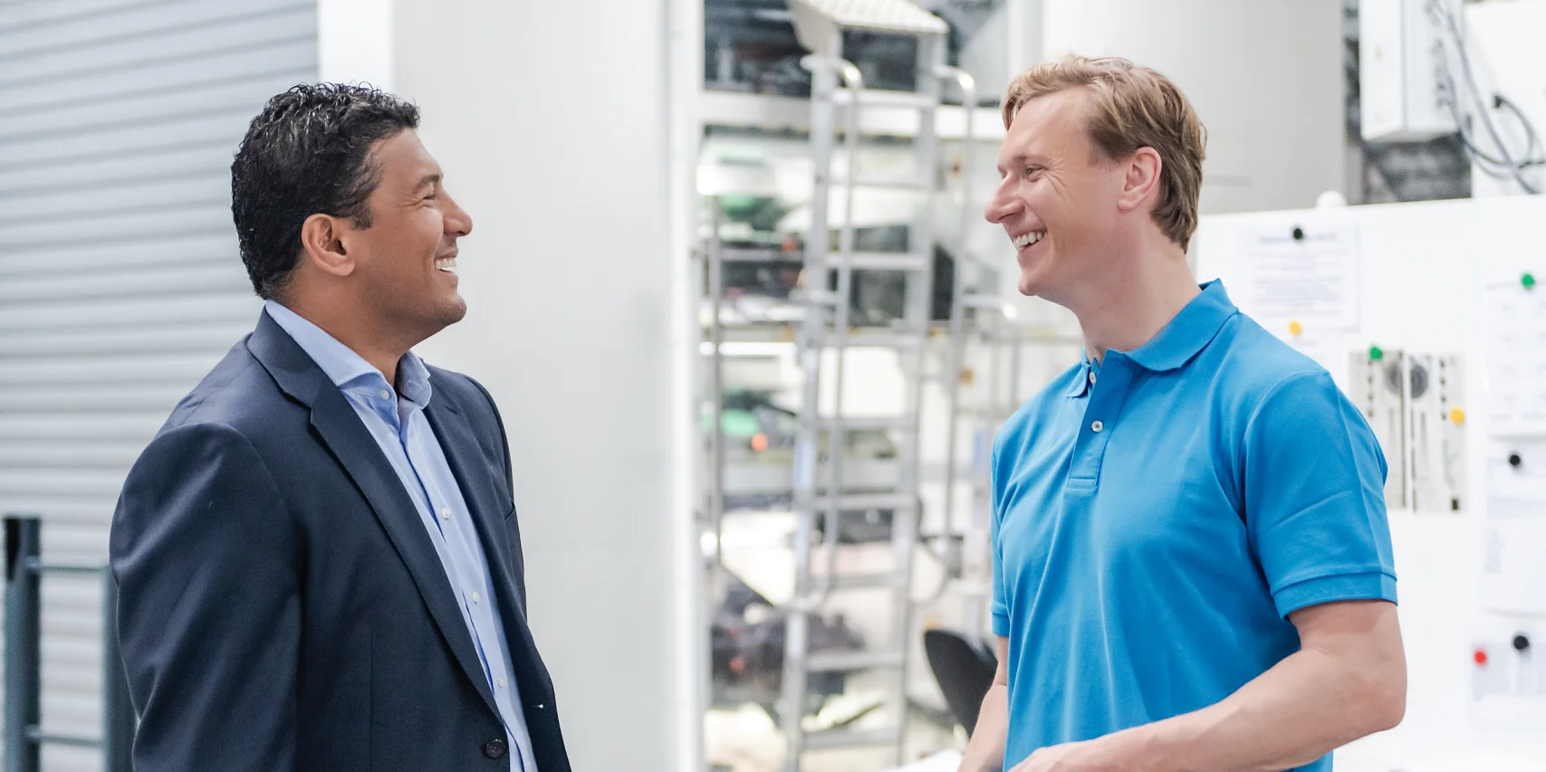 Two men are engaged in conversation in a modern industrial setting. The man on the left is dressed in a suit, while the man on the right wears a blue polo shirt and jeans. Equipment and machinery can be seen in the background. (This text has been generated by AI)