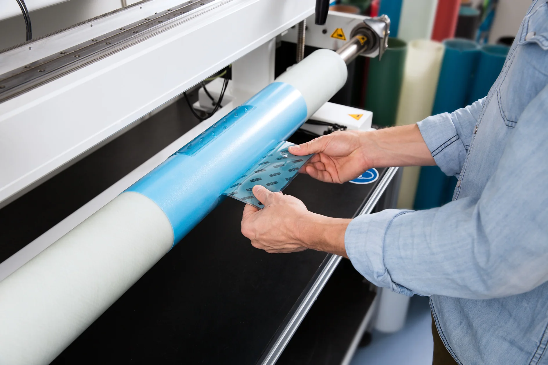 A person wearing a light denim shirt operates a machine with a large roll of blue and white material. They handle a transparent sheet with a pattern of shapes, possibly for printing or laminating. (This text has been generated by AI)
