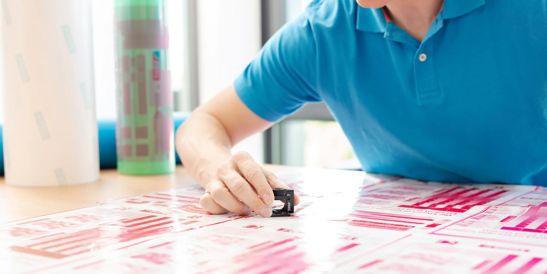 A person in a blue polo shirt examines printed material on a desk using a magnifying tool. Rolls of printed material are partially visible in the background. The print includes pink and red patterns and text, held together using tesa tape. (This text has been generated by AI)