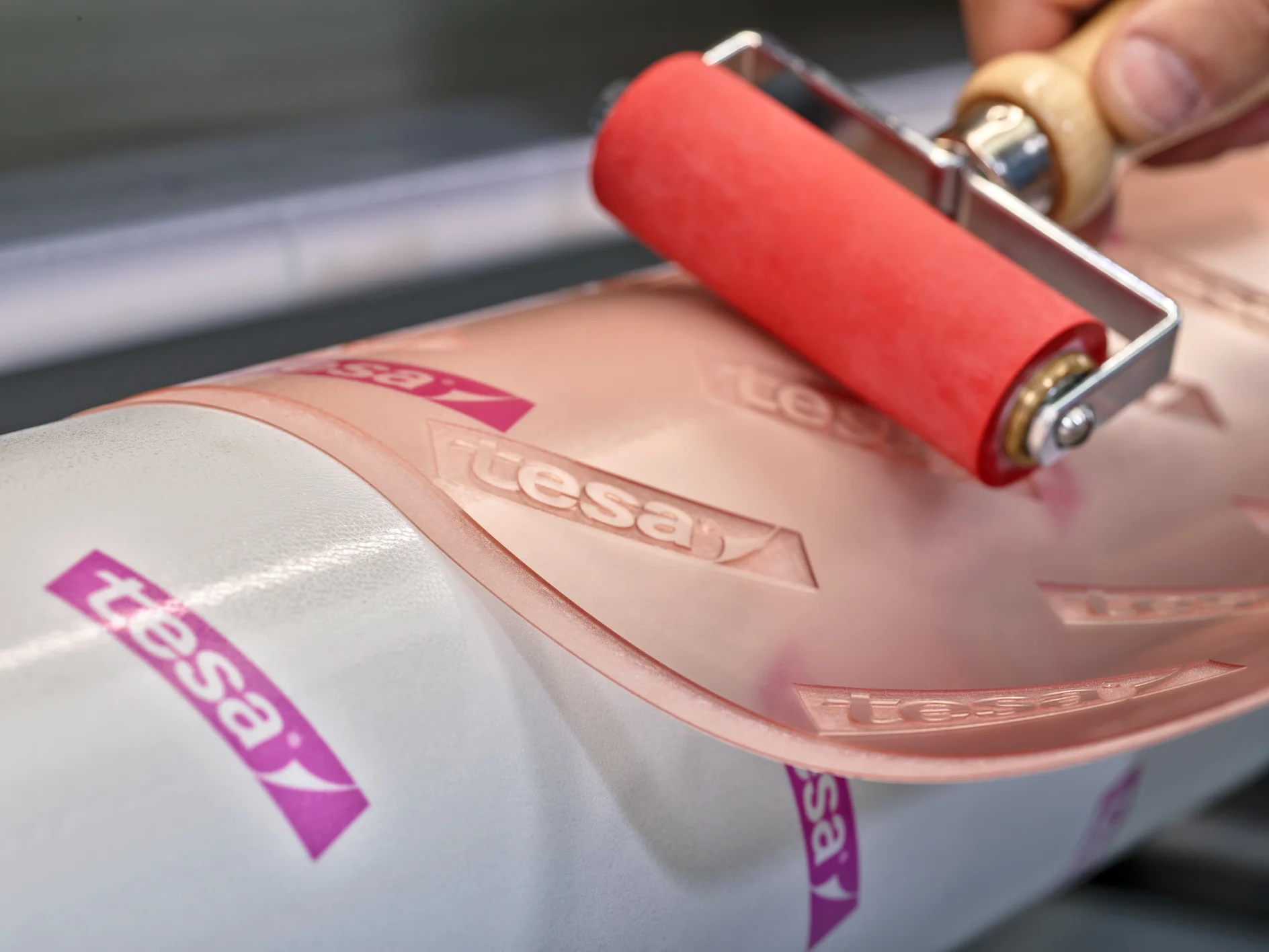 Close-up of a hand using a red roller on a clear adhesive film marked with the tesa tape logo. The film is being applied to a white cylindrical surface. (This text has been generated by AI)