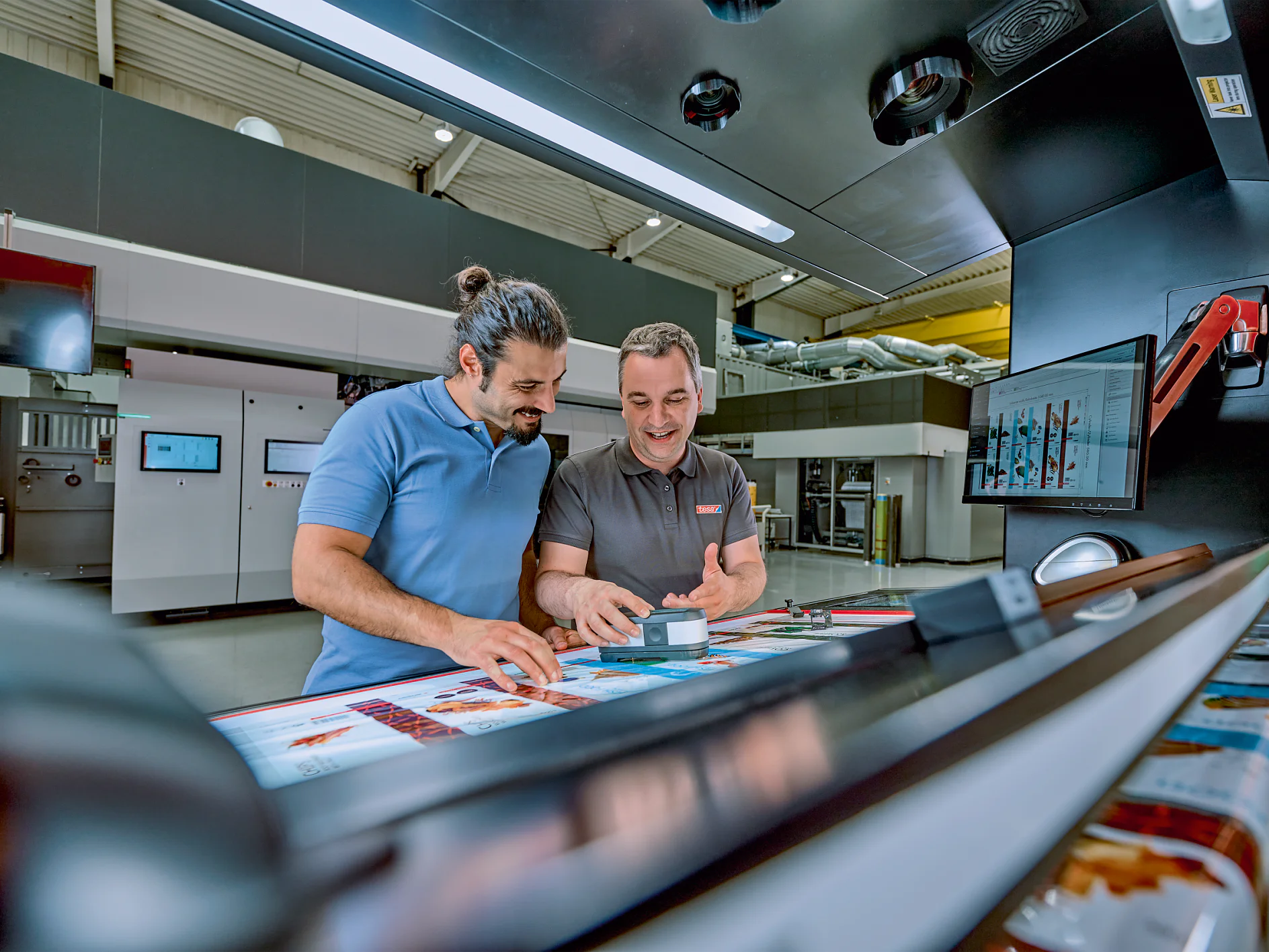 Two men are standing in a modern printing facility, examining colorful printed materials on a large machine. One man is holding a small box of tesa tape and smiling. There are screens and equipment in the background. (This text has been generated by AI)