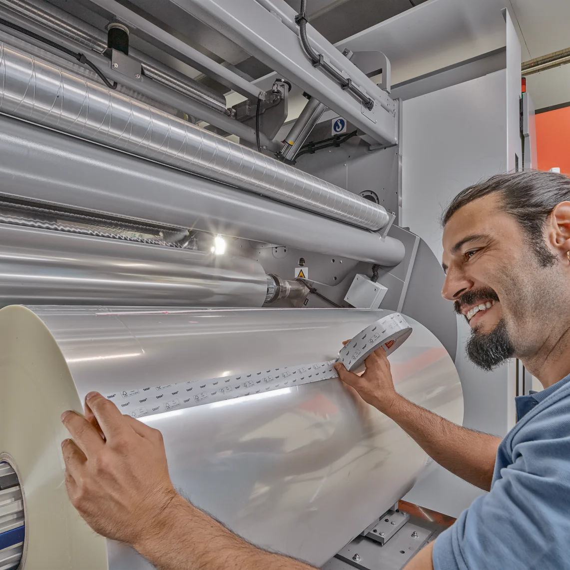 A man with a beard and a tied-back hairstyle operates a large industrial machine. He holds a strip of material running from a large roll of tesa tape. The machine and surroundings have a metallic and industrial appearance. (This text has been generated by AI)