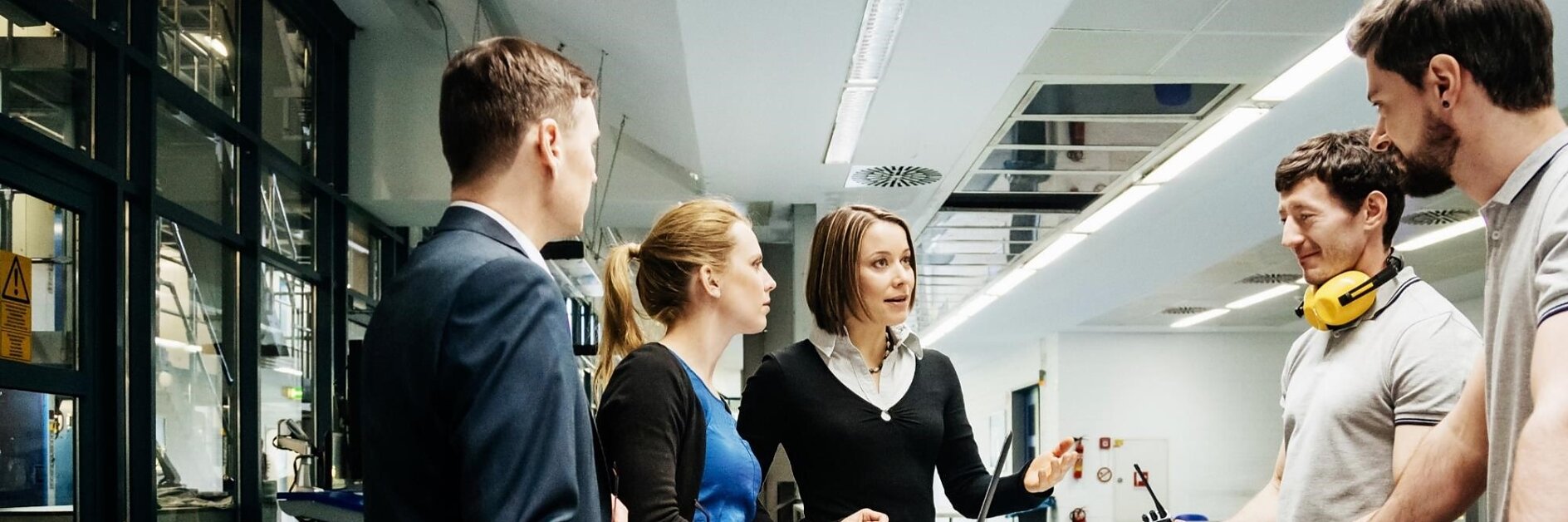 Five people are having a discussion around a desk in a modern office. One woman is gesturing while speaking, with tesa tape and a laptop in front of her. Two people are wearing casual clothing, and one is wearing yellow headphones around his neck.