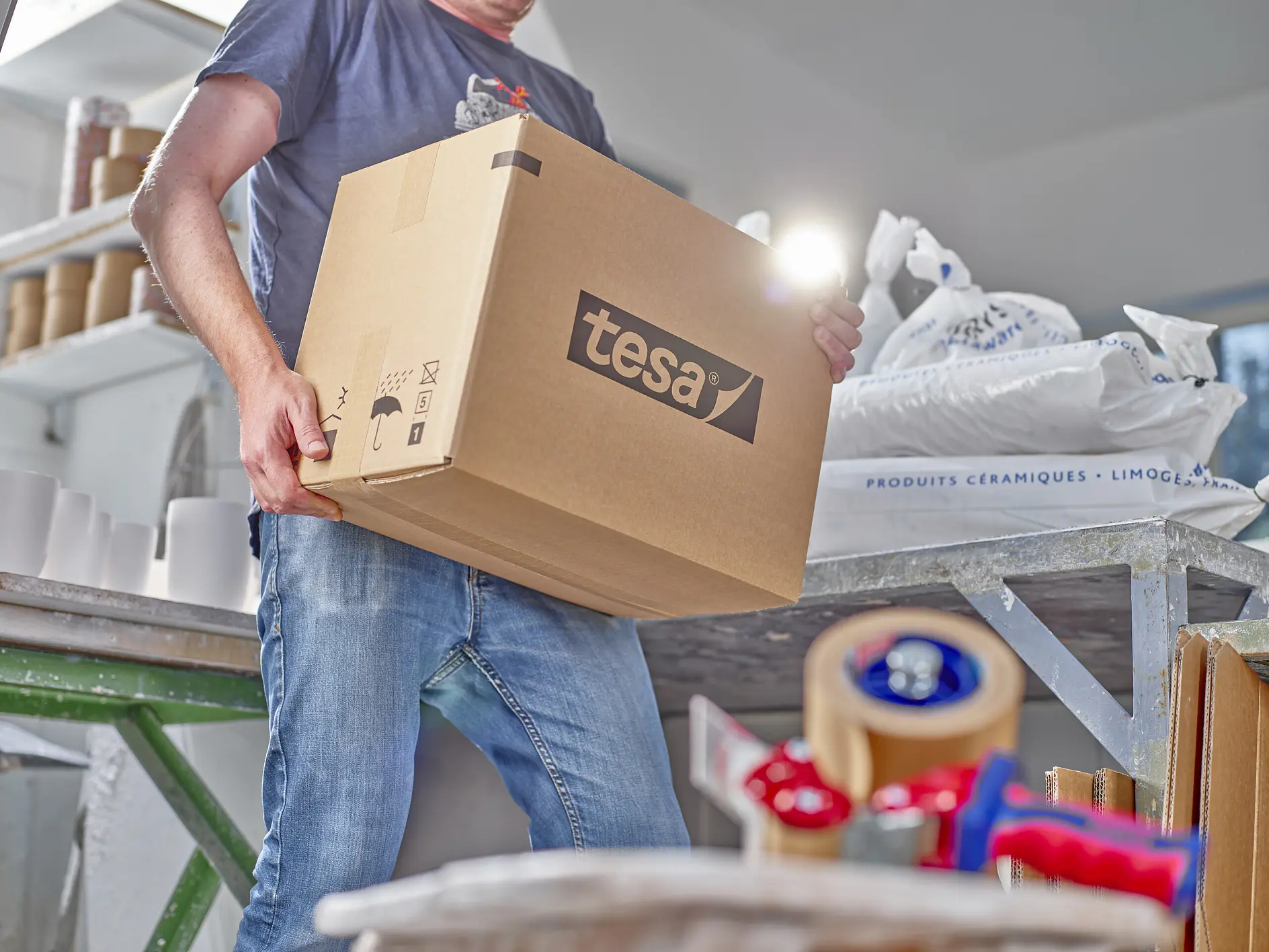A person holds a large cardboard box labeled tesa in a workspace. Various packing materials are scattered around, including tesa tape dispensers and white sacks. The person is wearing casual clothing, with jeans and a dark top. (This text has been generated by AI)