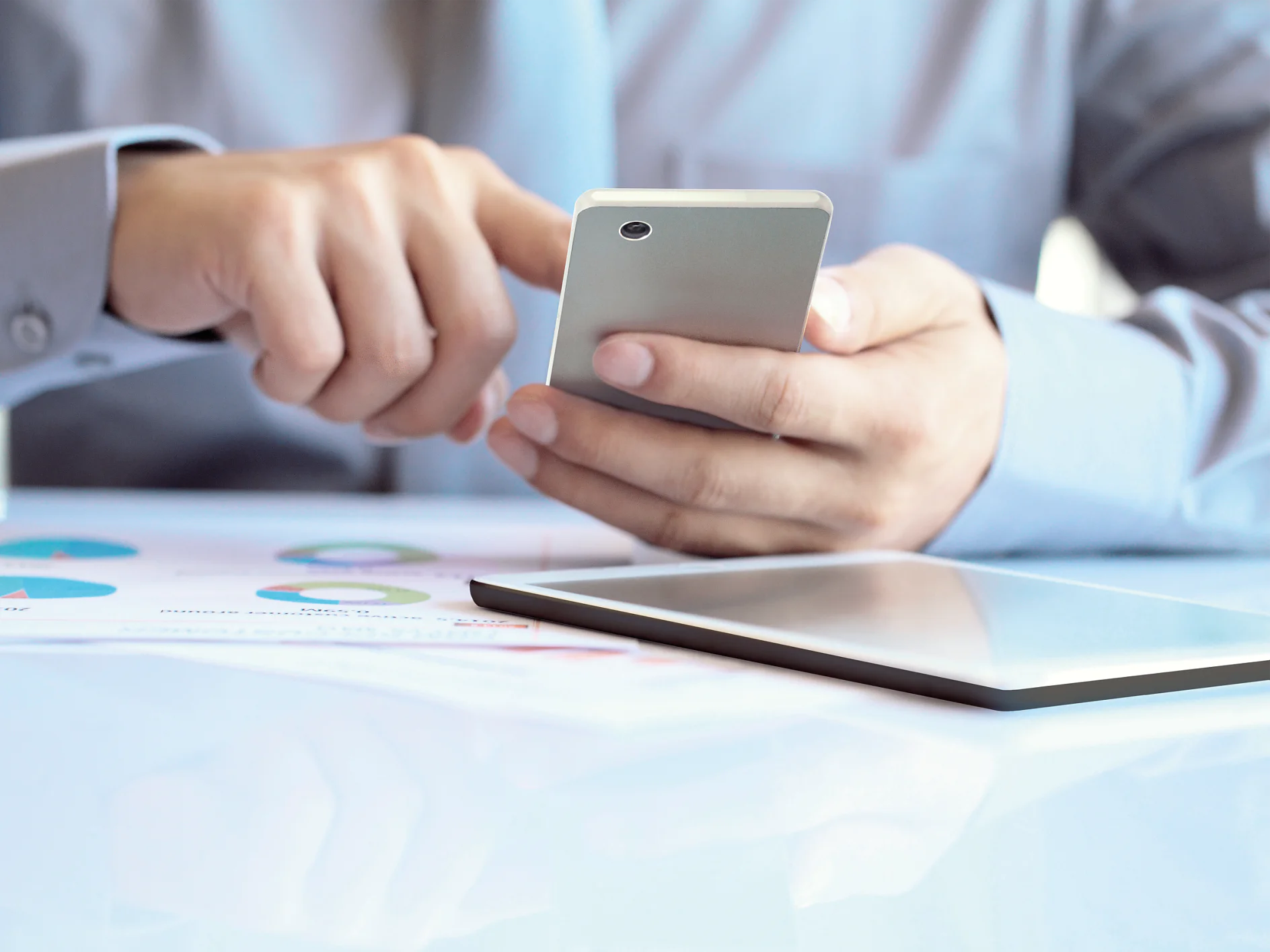 A person in a light blue shirt is using a smartphone, with an open binder or paper featuring colorful charts on the table, secured by tesa tape. Part of a tablet is visible in the foreground, slightly out of focus. The setting suggests a work environment. (This text has been generated by AI)
