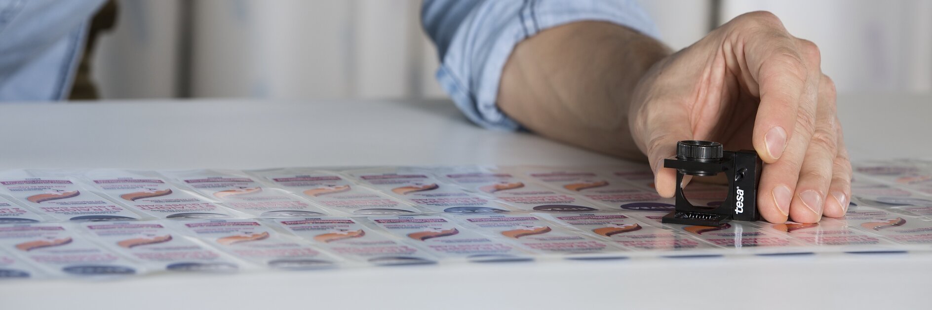 A person in a blue shirt examines a sheet of printed labels using a magnifying tool. The labels feature a colorful design with varying text. The hand is focusing on one particular label while holding the magnifying device and some tesa tape is visible on the nearby table.