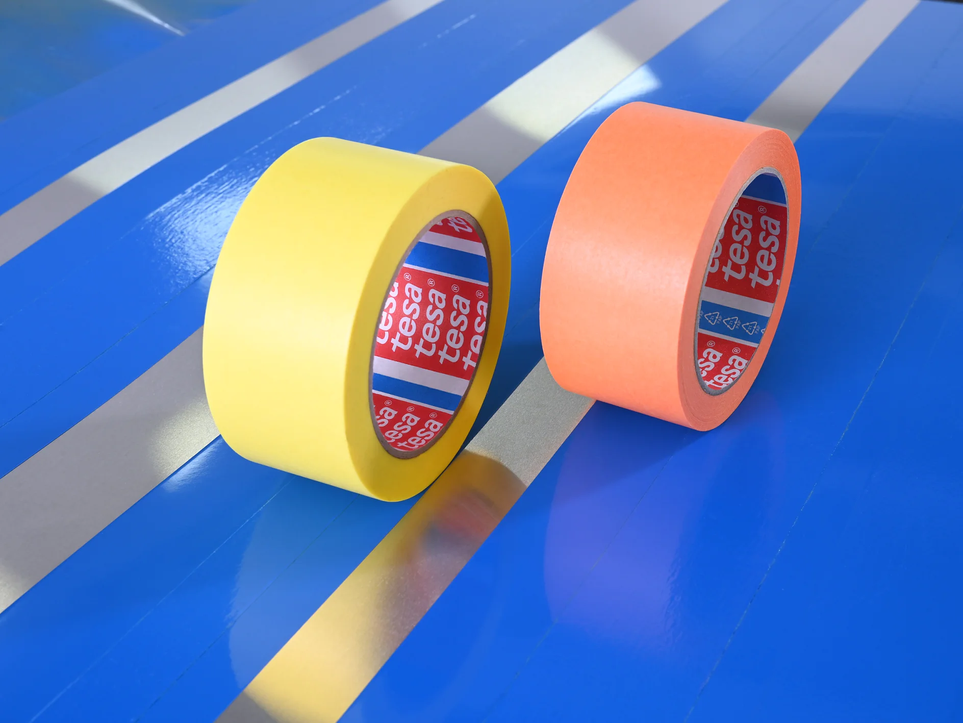 Two rolls of adhesive tesa tape on a blue and metallic-striped surface. One roll is yellow, and the other is orange. The inner labels display the brand name tesa. (This text has been generated by AI)