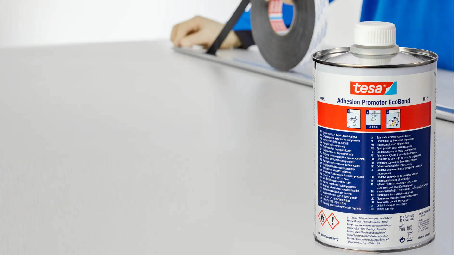 A person in a blue shirt wearing gloves holds a roll of tesa tape above a flat white surface. In the foreground, a can of tesa Adhesion Promoter EcoBond is visible with text and hazard symbols on its label. (This text has been generated by AI)