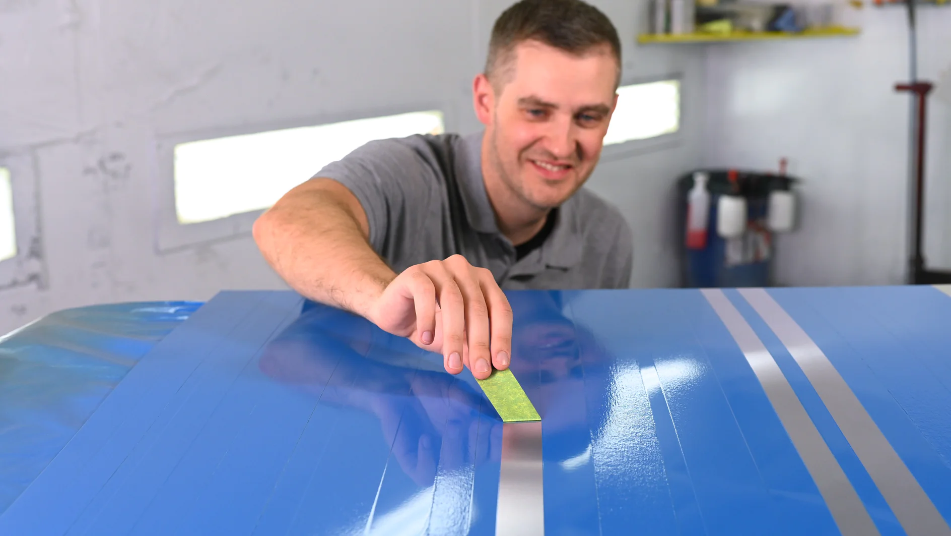 A person in a gray shirt applies a green strip of tesa tape to a blue surface with light gray stripes in a workshop. Shelves and equipment are visible in the background. (This text has been generated by AI)