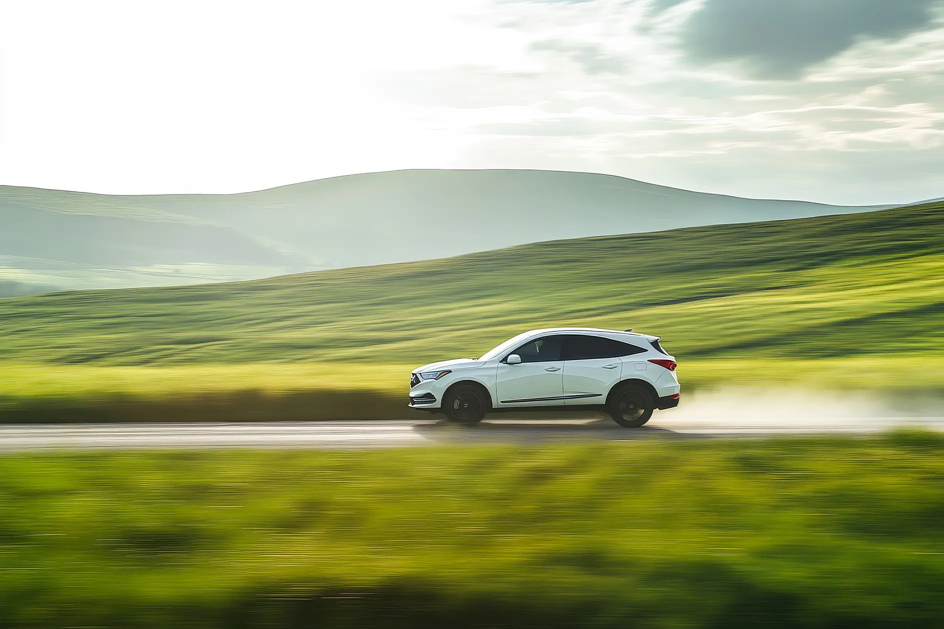 A white SUV drives quickly along a paved road through green hills under a partly cloudy sky, with tesa tape visible on its side.