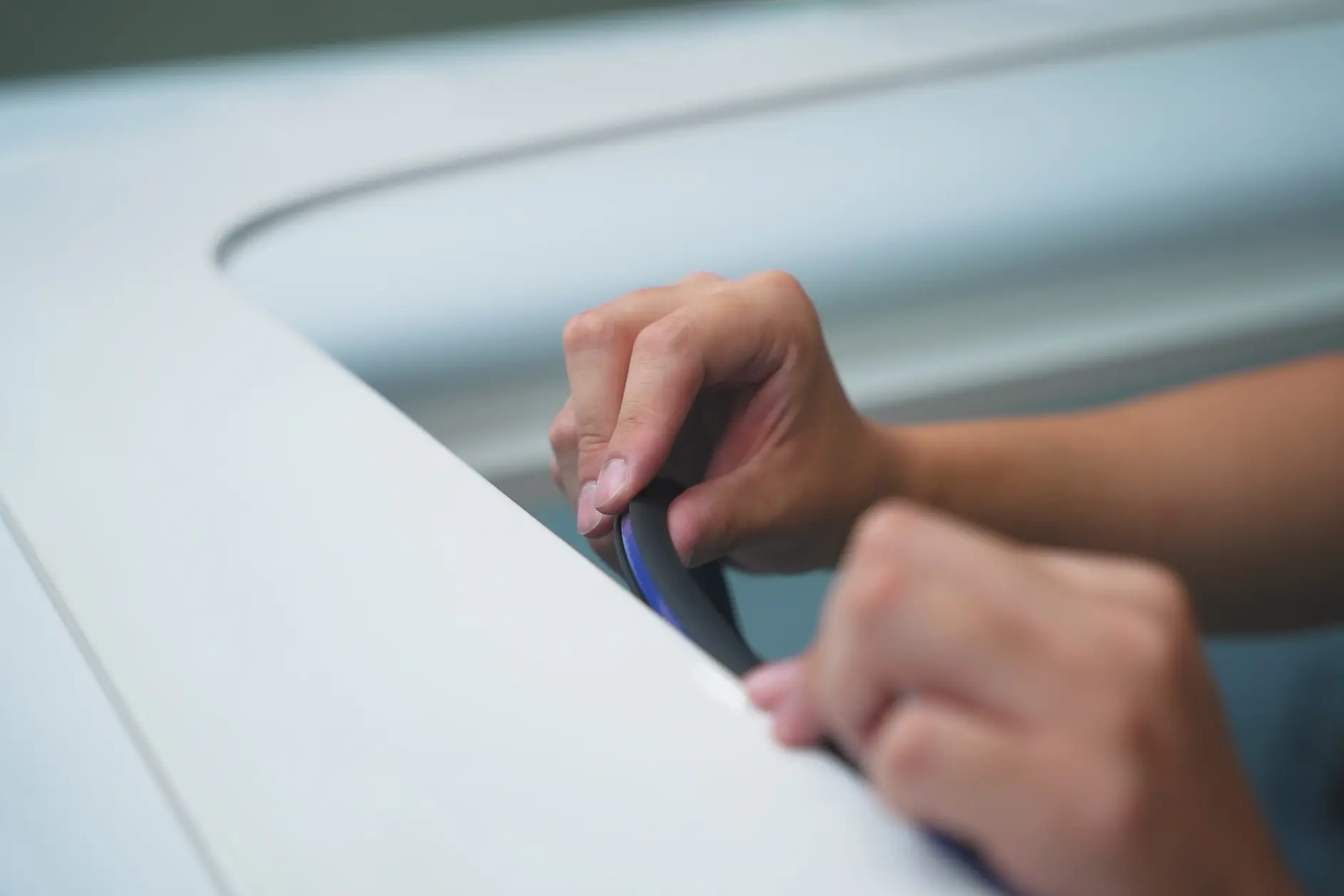 Close-up of two hands installing or adjusting a black rubber seal with tesa tape along the edge of a white surface, possibly a car.