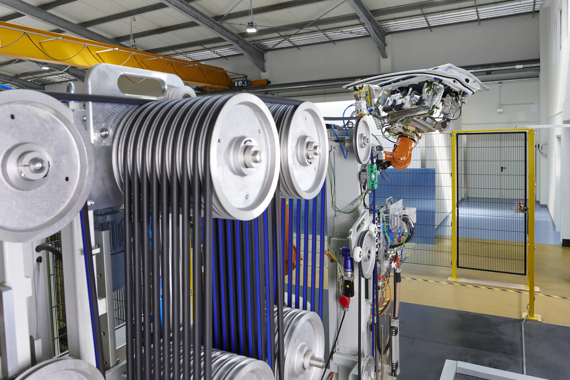 A close-up of industrial machinery in a factory, featuring metal pulleys with tesa tape on black belts, and a robotic arm to the right.