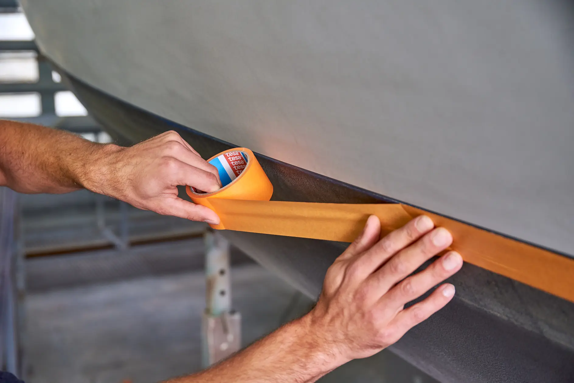 Hands applying orange tesa adhesive tape to the edge of a gray surface in an indoor workshop setting