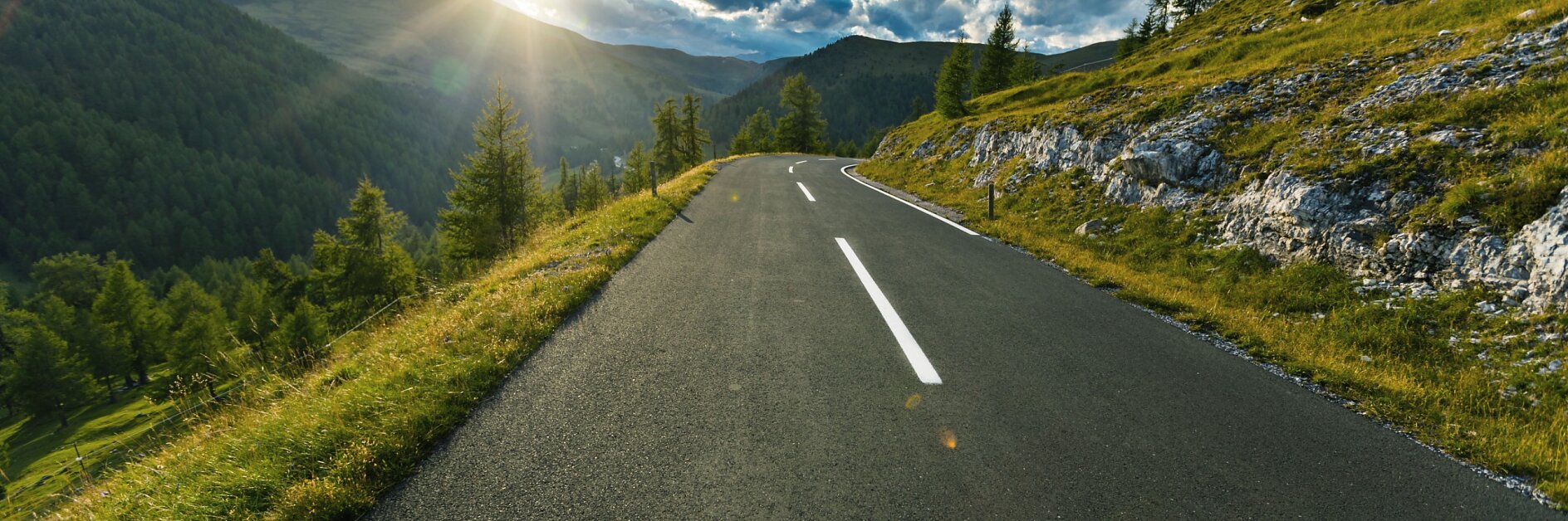 A paved road winds through green hills with trees and grass; the low sun casts long shadows beneath scattered clouds.