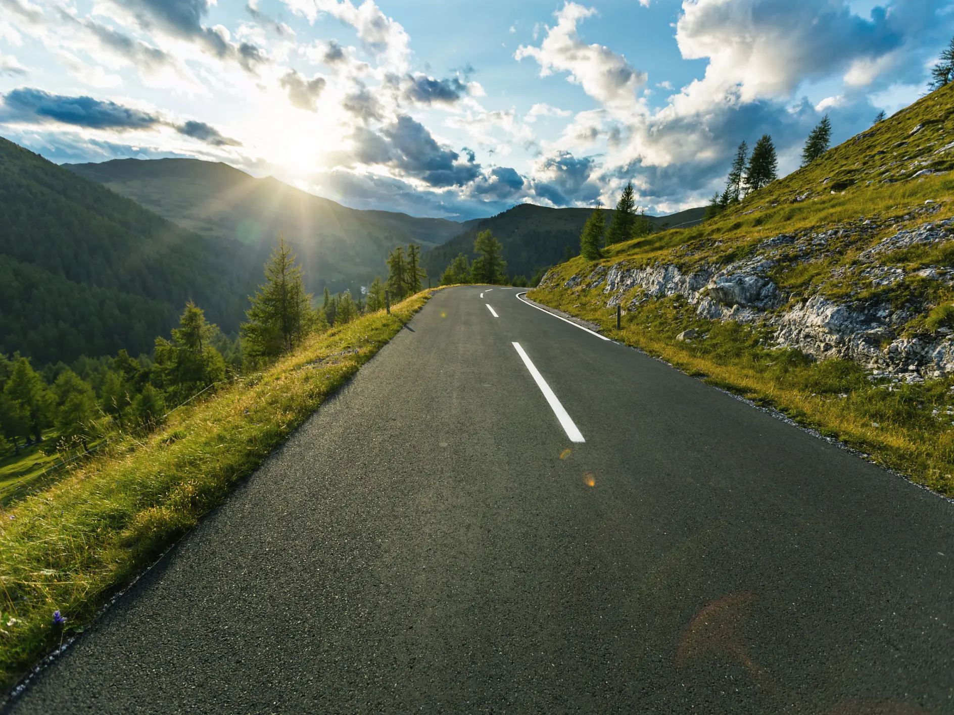 A paved road winds through green hills with trees and grass; the low sun casts long shadows beneath scattered clouds. (This text has been generated by AI)