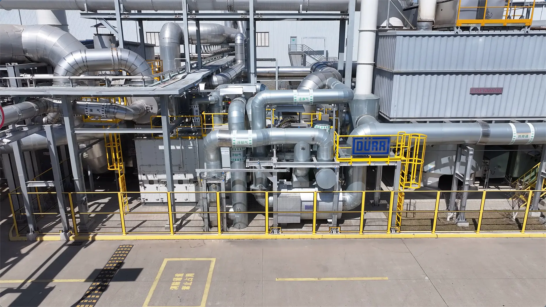 Industrial facility with metal pipes, ducts, equipment, yellow safety railings and walkways. Dürr sign on machinery. tesa tape visible.