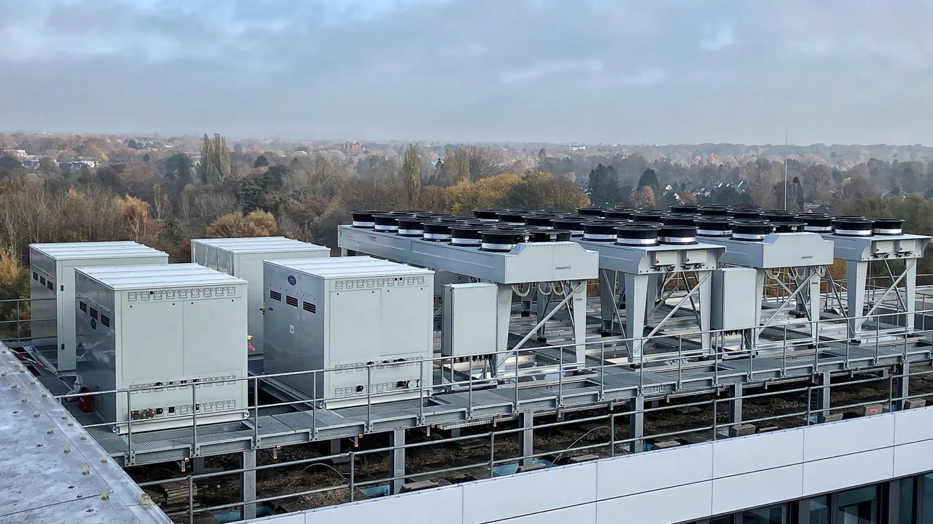 Industrial HVAC and cooling systems sit on a rooftop, overlooking trees and distant buildings under a partly cloudy sky. (This text has been generated by AI)