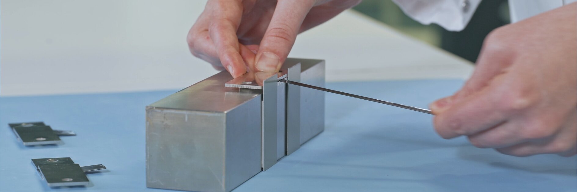 A person in a white lab coat uses a thin metal tool on a rectangular metallic device placed on a blue surface. Two additional metal pieces lie nearby, secured with tesa tape. The scene appears to be in a laboratory setting.
