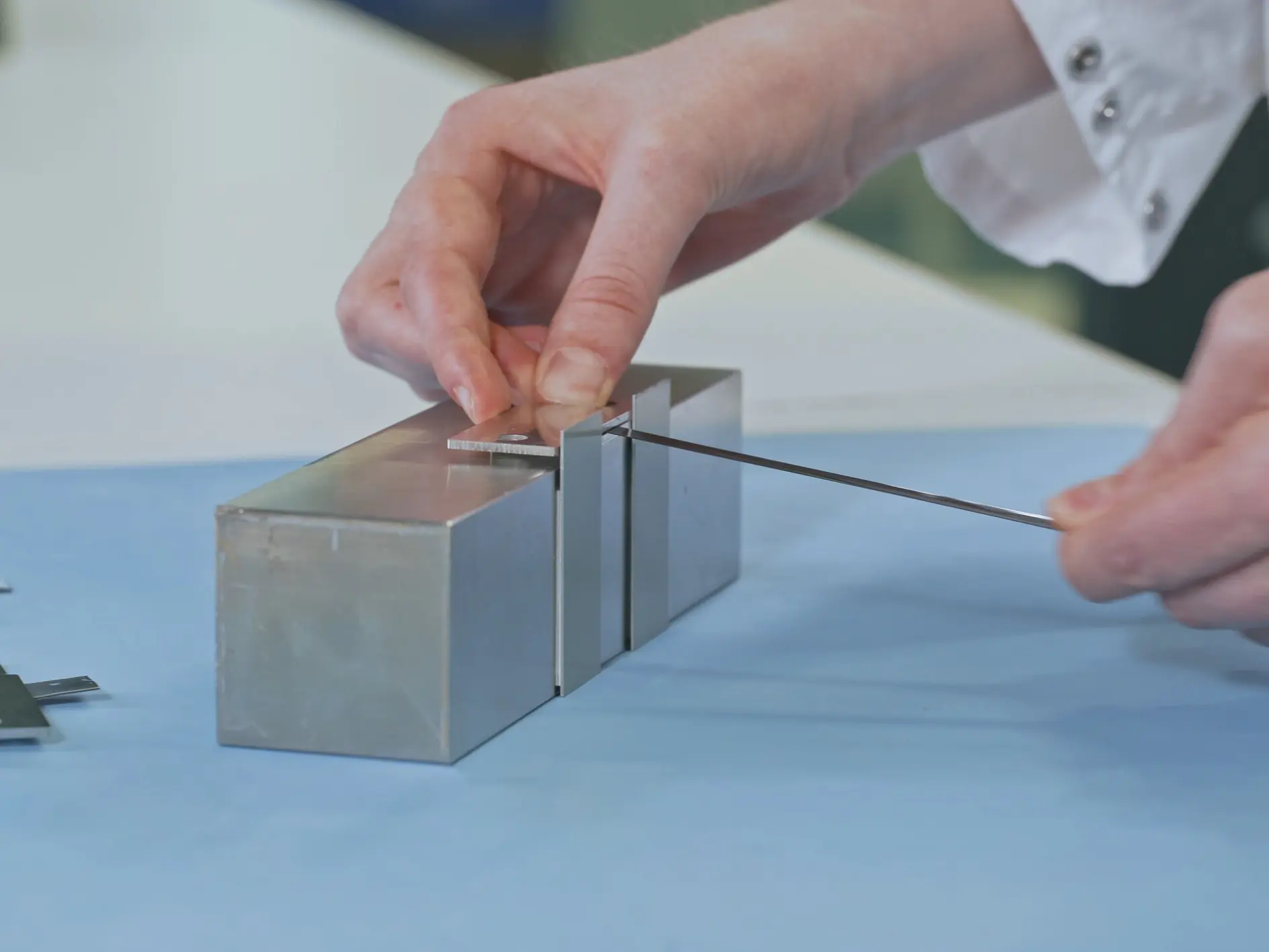 A person in a white lab coat uses a thin metal tool on a rectangular metallic device placed on a blue surface. Two additional metal pieces lie nearby, secured with tesa tape. The scene appears to be in a laboratory setting. (This text has been generated by AI)