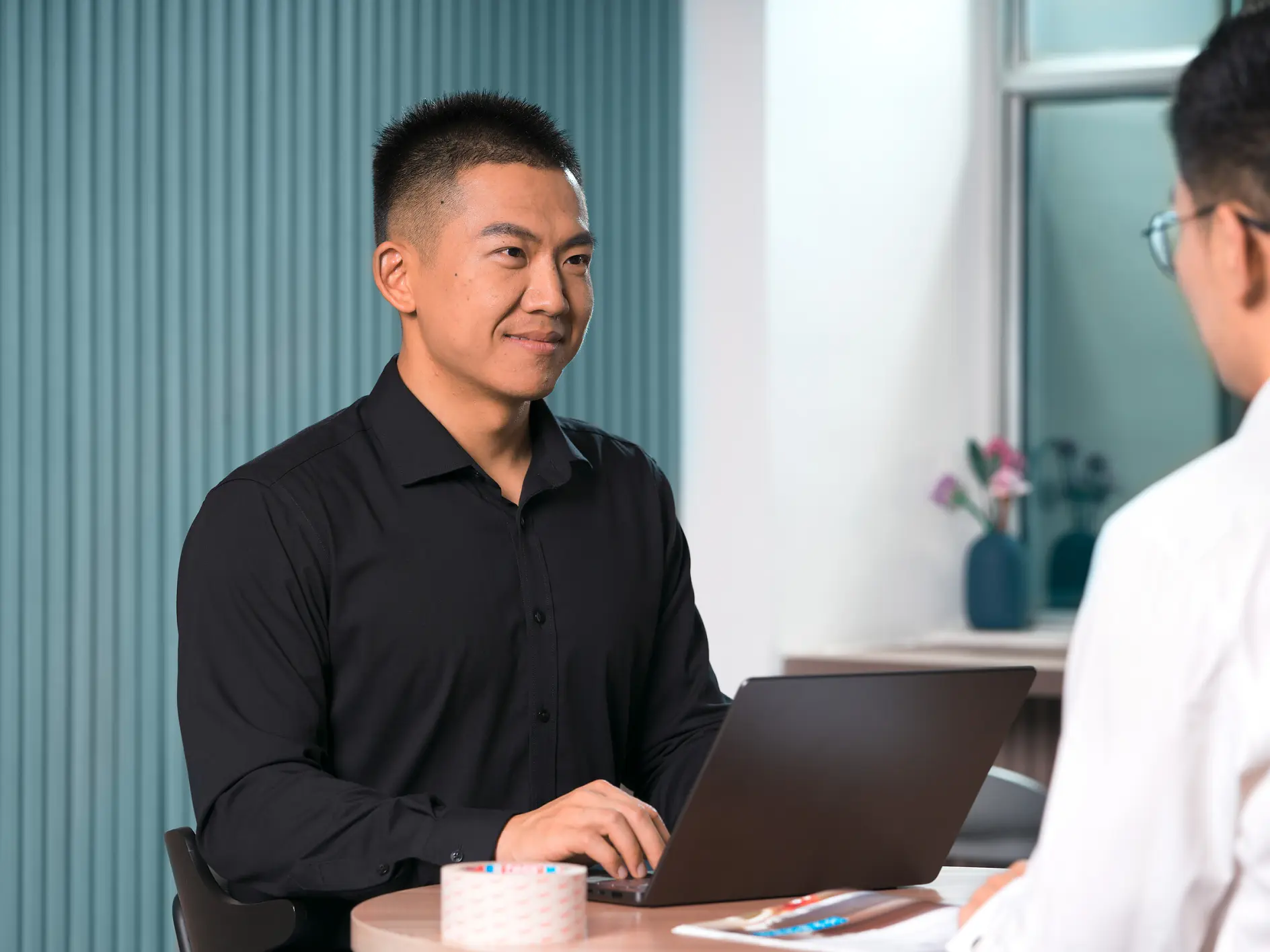Two men in an office: one in a black shirt smiles while working on a laptop, while the other, seen from behind in a white shirt, listens attentively. (This text has been generated by AI)
