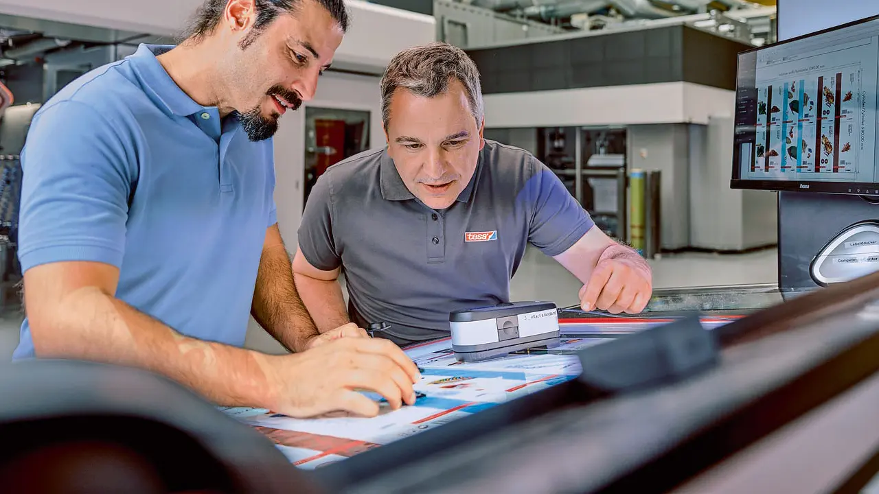 Two men are examining printed pages under a light in an industrial setting. One holds a magnifying glass, while the other points at the paper secured with tesa tape. A computer monitor displaying graphics is visible on the side. Both are wearing casual shirts.