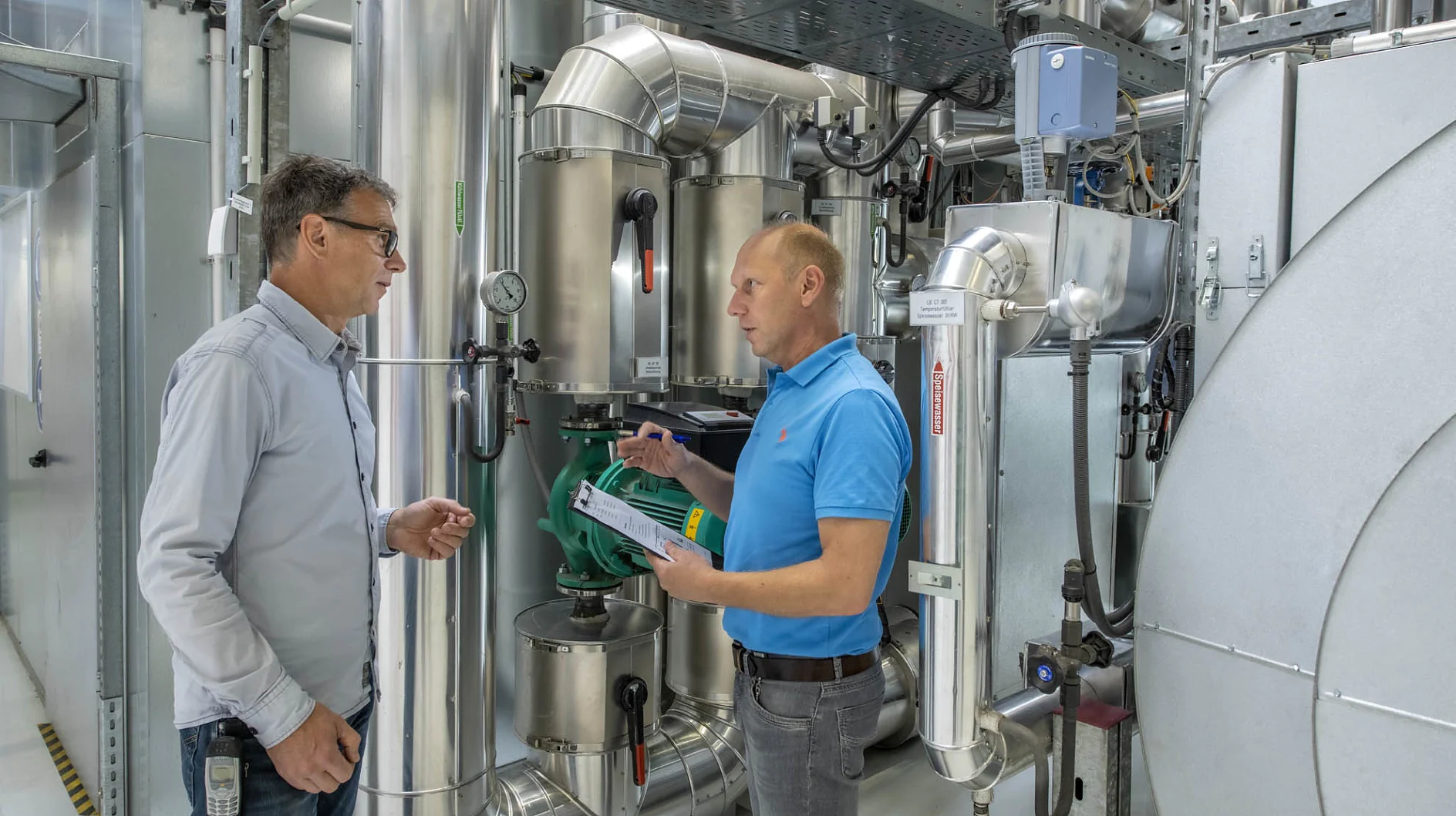 In an industrial setting surrounded by metal pipes and machinery, two men stand engaged in discussion. One man holds a clipboard and gestures as the other listens attentively. Both are dressed in casual clothes. They focus on using tesa tape for their tasks and projects, confident in its quality and performance. (This text has been generated by AI)