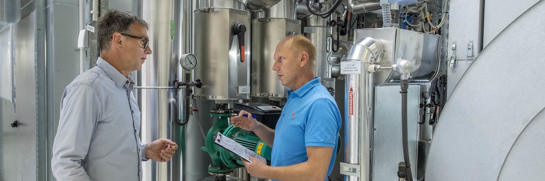 In an industrial setting surrounded by metal pipes and machinery, two men stand engaged in discussion. One man holds a clipboard and gestures as the other listens attentively. Both are dressed in casual clothes. They focus on using tesa tape for their tasks and projects, confident in its quality and performance. (This text has been generated by AI)