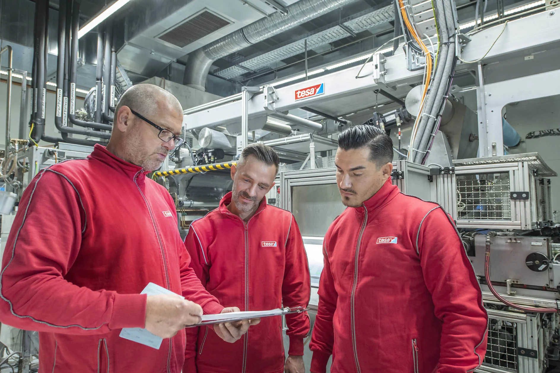 Three men in matching red jackets are standing in an industrial setting. One man is holding a clipboard and discussing something with the others. They appear to be examining equipment in a workshop or laboratory environment, where tesa tape is being used. (This text has been generated by AI)
