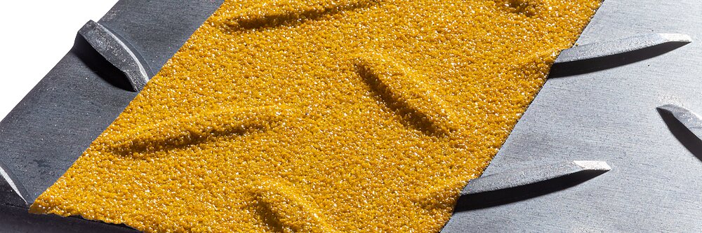 Close-up of a textured surface containing a yellow, gritty area with diagonal ridges, bordered by sections of dark gray metal with parallel grooves. The contrast highlights the different materials and textures.