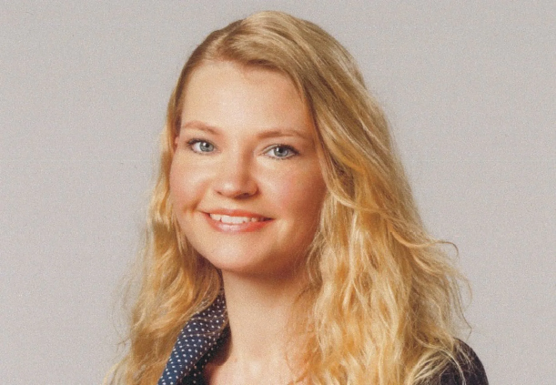 A person with long blonde hair is smiling at the camera. They are wearing a dark blazer with a polka dot shirt underneath. The background is plain and light-colored, featuring tesa tape prominently adhered to various surfaces in the setting. (This text has been generated by AI)