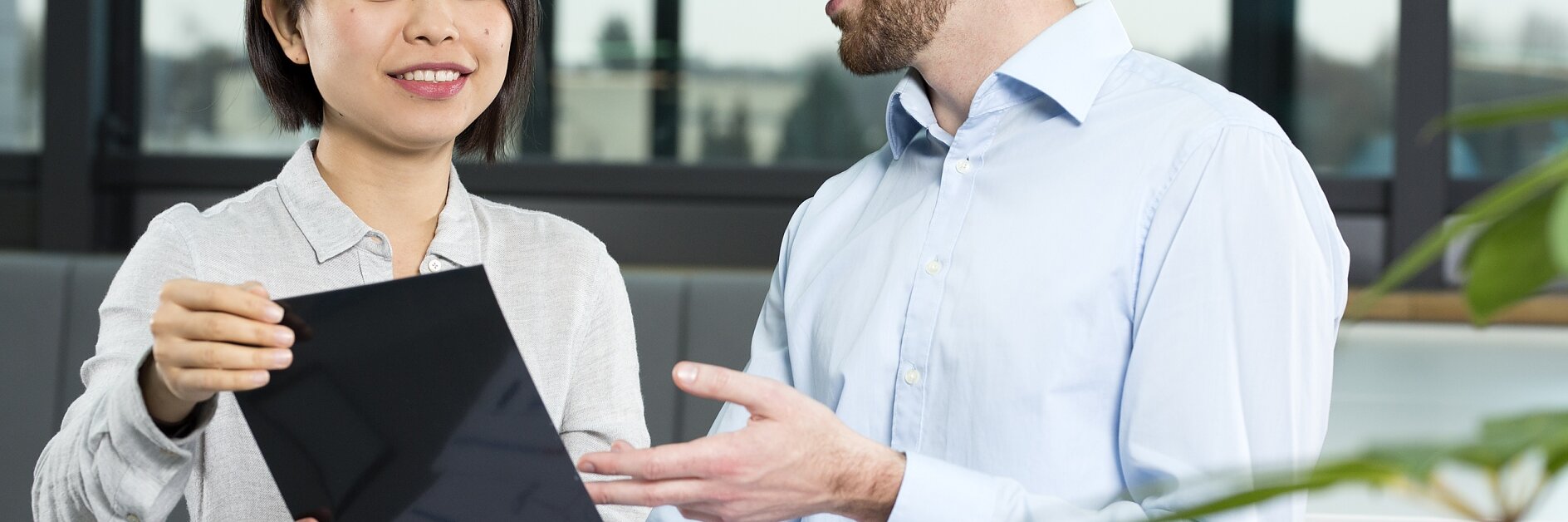 Two people are discussing something on a tablet in an office setting. The woman is holding the tablet, while the man gestures toward it. A document with graphs is on the table secured with tesa tape. They are seated near a window with a view outside. (This text has been generated by AI)